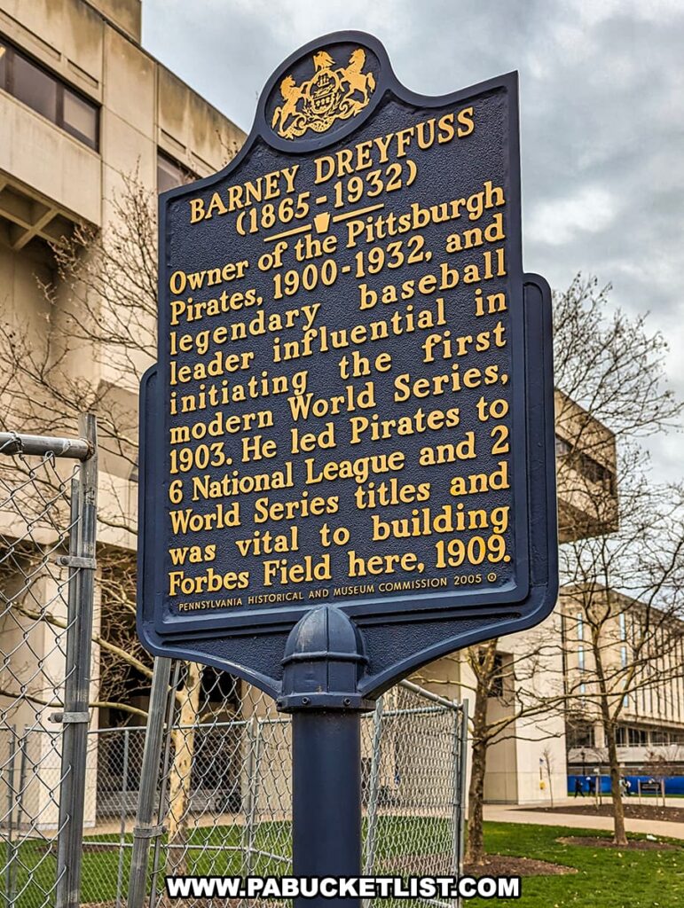 A historical marker commemorating Barney Dreyfuss (1865-1932), the owner of the Pittsburgh Pirates from 1900 to 1932, is mounted on a metal post and situated in front of a chain-link fence with a building in the background. The marker, with raised golden lettering on a navy blue background, credits Dreyfuss as a legendary baseball leader who was influential in initiating the first modern World Series. It notes that he led the Pirates to 6 National League titles and 2 World Series titles, emphasizing his importance in the construction of Forbes Field in 1909. The marker is designated by the Pennsylvania Historical and Museum Commission, dated 2005.