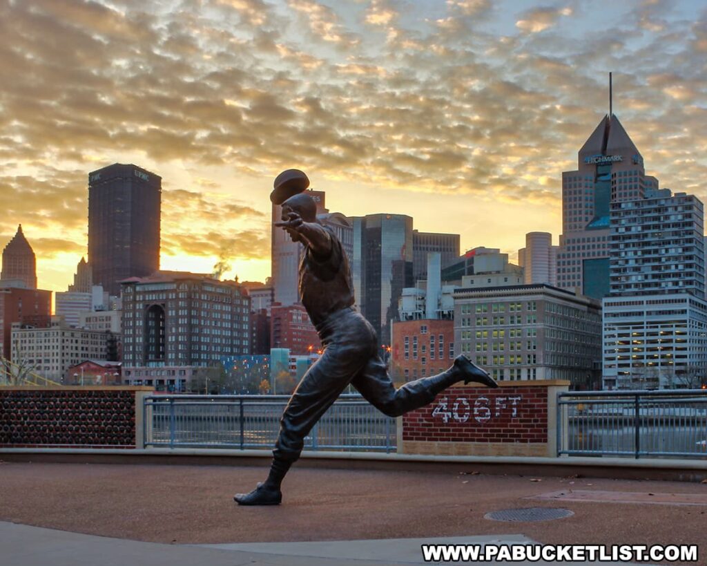 Silhouetted against a striking sunrise sky with scattered clouds, a bronze statue captures the dynamic motion of baseball player Bill Mazeroski in mid-run, right arm trailing and left leg lifted high. The player is depicted wearing a period uniform and cap, suggesting a connection to the historic era of Forbes Field. In the background, the Pittsburgh skyline rises, blending modern buildings with the timeless spirit of the game. The statue is poised in front of a red brick wall that bears the distance marker "406 FT," echoing the outfield wall distance at Forbes Field.