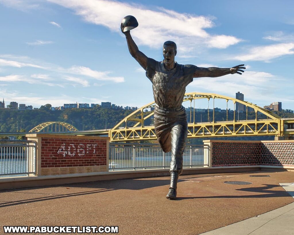 A bronze statue of baseball player Bill Mazeroski in mid-stride, celebrating a moment of triumph with his right arm extended upwards holding a baseball cap, is positioned prominently in the foreground. Behind the statue is a red brick wall with the distance marker "406 FT" signifying the depth to center field, reminiscent of the measurements at Forbes Field. The picturesque backdrop features the Allegheny River and one of Pittsburgh's iconic yellow bridges, with the cityscape and hills in the distance. The scene is bathed in the warm glow of afternoon sunlight, evoking the historic connection to Forbes Field, the former home of the Pittsburgh Pirates.