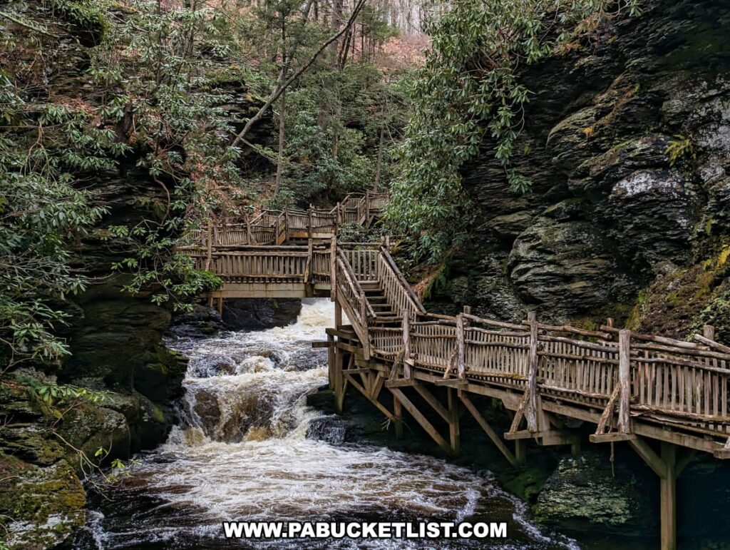 A serene view of Little Bushkill Creek as it flows through a rocky gorge at Bushkill Falls in Pike County, Pennsylvania. A wooden boardwalk with a rustic railing crosses the creek, connecting two platforms that provide visitors with vantage points to enjoy the surrounding natural beauty. The boardwalk is nestled among the towering cliffs and lush greenery of rhododendrons, typical of the Pocono Mountains region. The tranquil yet dynamic scene captures the essence of Bushkill Falls, known as the "Niagara of Pennsylvania," with its well-preserved environment and accessible paths that invite exploration and appreciation of the area's scenic waterfalls and woodlands.