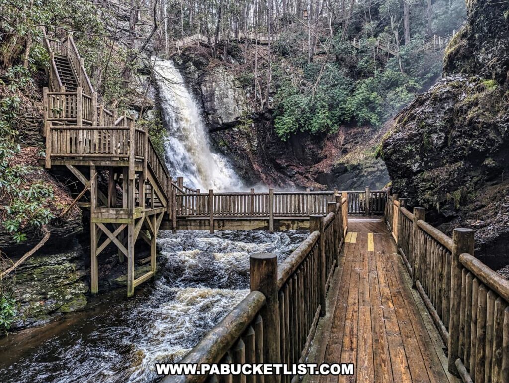 The scene is set at Bushkill Falls in Pike County, Pennsylvania, where a wooden observation deck provides a close-up view of one of the park's prominent waterfalls. The deck, part of a comprehensive network of well-maintained boardwalks, extends over a turbulent stream, leading visitors toward a two-tiered viewing platform. This structure sits adjacent to the waterfall, allowing visitors to feel the mist and hear the thunderous sound of the water as it crashes into the basin below. Lush greenery and mossy rocks frame the waterfall, highlighting the natural beauty and forested environment of the Pocono Mountains.