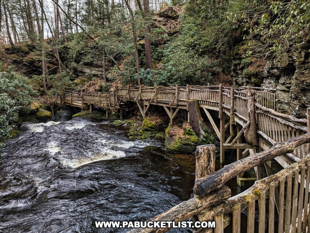 A sturdy wooden boardwalk at Bushkill Falls in Pike County, Pennsylvania, winds its way through a lush landscape. It crosses over a lively, dark stream that runs rapidly beneath, frothing as it interacts with the natural rocky barriers. The railings of the boardwalk, coupled with the moss-covered boulders and the mature, verdant trees that surround it, create an immersive outdoor experience.