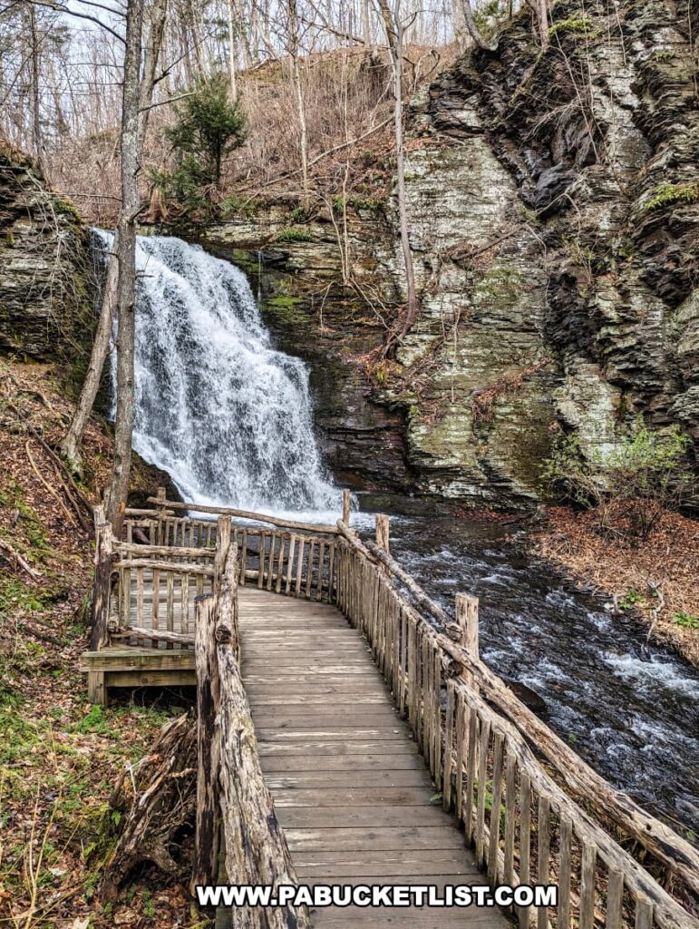 A wooden boardwalk leads visitors up close to the dynamic Bridal Veil Falls at Bushkill Falls in Pike County, Pennsylvania. The boardwalk, with its rustic railings, allows for a safe and immersive viewing experience of the waterfall's powerful cascade. Water rushes down over mossy rocks and into a lively stream that runs alongside the path. The scene is framed by the striated rock face on one side and a variety of trees, some bare and others evergreen, on the other, depicting a cool, late autumn or early spring day in the Pocono Mountains.
