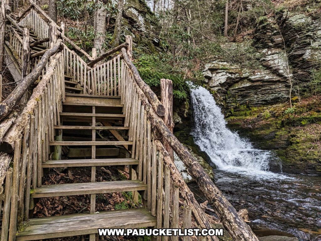 A rustic wooden staircase, part of the extensive boardwalk system at Bushkill Falls in Pike County, Pennsylvania, descends through a forested area towards a viewing platform near the Bridesmaid Falls. The handcrafted railings of the stairs, composed of branches and timber, lead the eye downwards toward the waterfall, which gushes energetically into a rock-strewn stream below. The falls are set against a backdrop of layered shale and sandstone, typical of the geological formations in the Pocono Mountains. Overhead, the canopy is sparsely leaved, as it is early spring pictured in the photo.