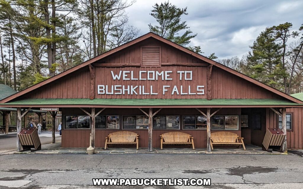 The entrance to Bushkill Falls, located in Pike County, Pennsylvania, is marked by a welcoming visitor center with a dark wooden façade and a gabled roof. Large white letters on the roof spell out "Welcome to Bushkill Falls," greeting guests as they arrive. In front of the building, wooden benches offer a place for visitors to rest. The center appears to house the souvenir shop, as indicated by the sign above the entrance, and provides amenities and information for those looking to explore the "Niagara of Pennsylvania" and its network of trails and scenic waterfalls within the beautiful Pocono Mountains.