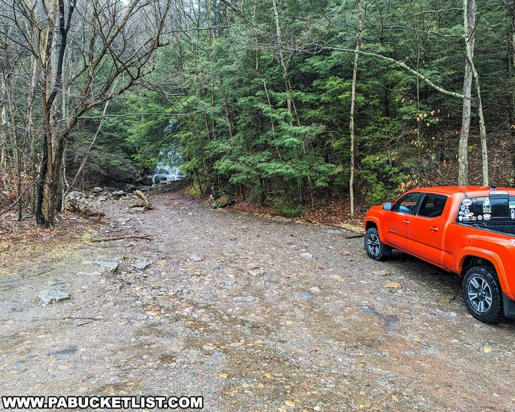 A vibrant orange pickup truck is parked in a gravel lot surrounded by a mix of evergreen and leafless deciduous trees, indicative of the changing seasons. In the background, the natural beauty of Buttermilk Falls in the Bear Creek Preserve of Luzerne County, Pennsylvania, can be glimpsed through the woodland, inviting exploration and adventure in this serene setting.