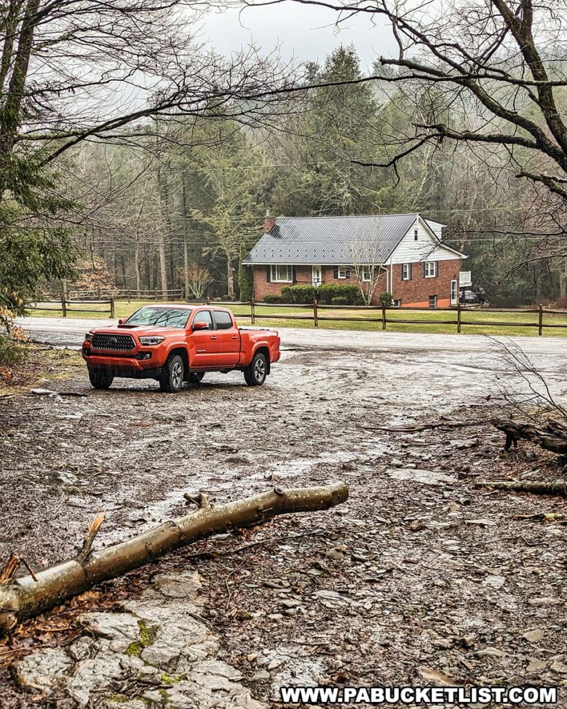 An orange pickup truck is parked on a gravel lot near Buttermilk Falls within the Bear Creek Preserve, Luzerne County, Pennsylvania. The vehicle stands out against a backdrop of a misty, forested landscape and a classic brick house just across the quiet country road. A fallen branch in the foreground adds a rustic touch to this peaceful rural setting.