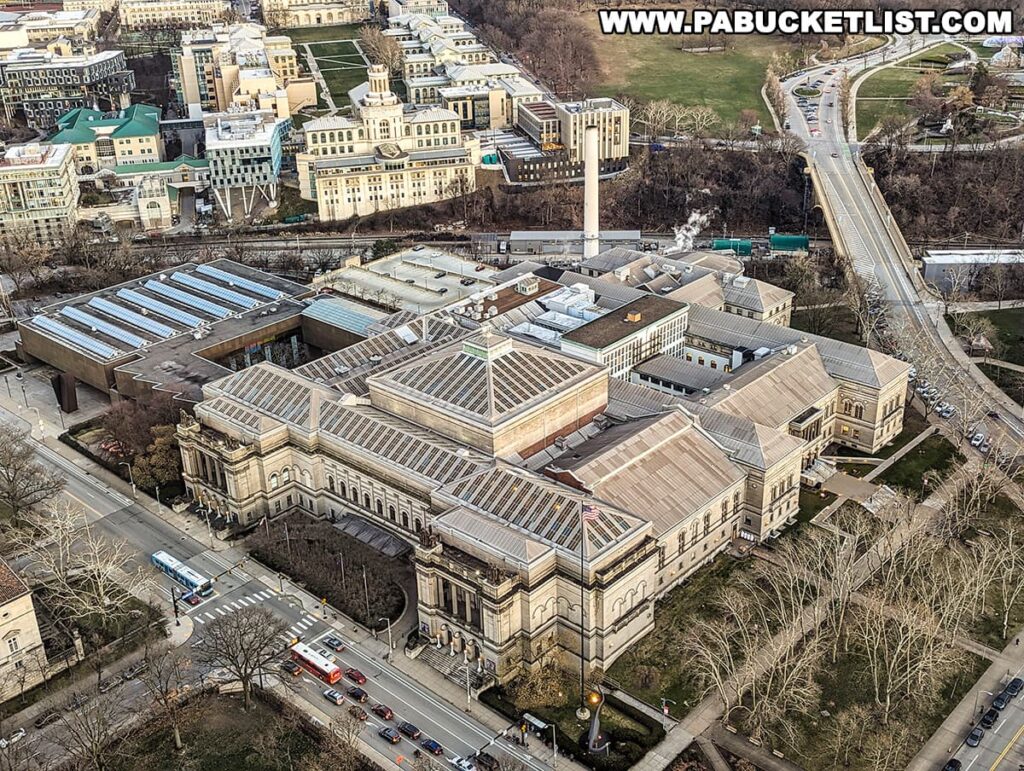An aerial view of the Carnegie Museums of Art and Natural History in the Oakland neighborhood of Pittsburgh, Pennsylvania. The grand, stone edifices of the museum complex are seen amidst the urban landscape, with expansive glass skylights and classic architectural details. Nearby, the intricate layout of university buildings and green spaces suggests the proximity to a campus environment. Traffic is visible on the surrounding streets, and a park area with bare trees indicates the photo was taken during the cooler months. The overall scene captures the cultural and academic heartbeat of the area once associated with Forbes Field and its historical significance to Pittsburgh.