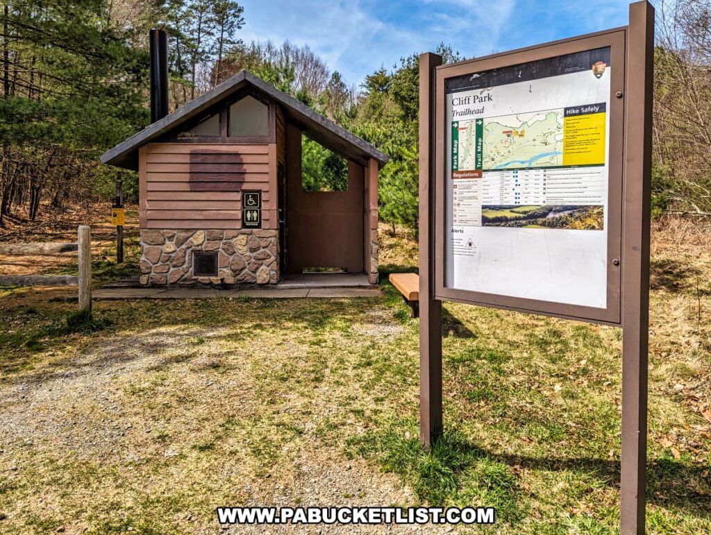 The trailhead of Cliff Park in Pike County, Pennsylvania, featuring a small restroom with a stone base and is adorned with accessibility symbols. The signboard displays a map of the trail system, safety information, regulations, and alerts for hikers. In the background, a forest with tall pine trees and a clear sky suggests a serene natural setting for the start of the trails that lead to scenic overlooks above the Delaware River. A wooden bench sits next to the signboard, offering a place to rest or prepare for the hike.