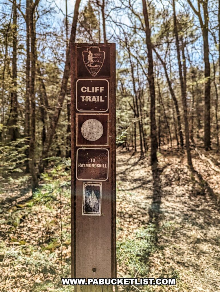 A wooden signpost on the Cliff Trail in Pike County, Pennsylvania, with directional markers against a backdrop of a wooded area. The signpost features the Pennsylvania state outline with the National Park Service logo at the top, followed by a black and white sign reading "CLIFF TRAIL", and below that, a directional sign pointing "TO RAYMONDSKILL". A white circular blaze on the post indicates the trail route. The trees surrounding the signpost are mostly bare, suggesting it's either late fall or early spring. The ground is covered with a carpet of brown leaves, and the forest has a quiet, serene atmosphere.
