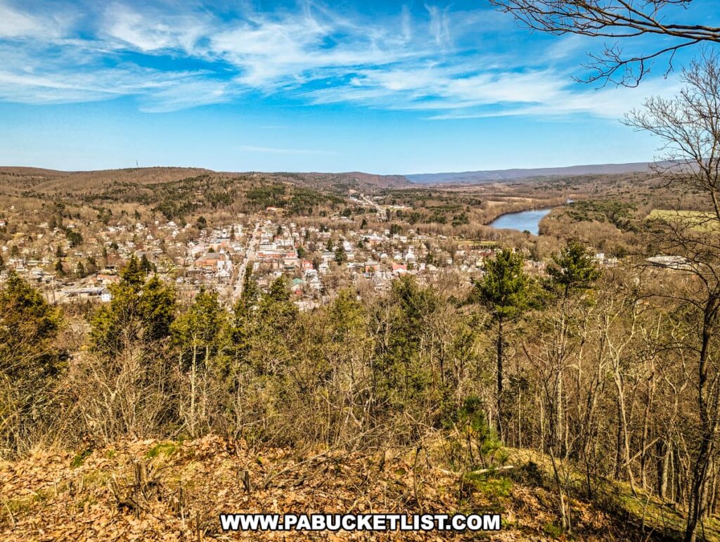 A view from the Cliff Trail in Pike County, Pennsylvania, captures Milford spread out amidst a forested landscape. Buildings and homes in various colors and sizes are neatly arrayed in the valley, with streets intersecting at regular intervals. Beyond the town, the Delaware River curves gracefully between the hills. Trees with early spring foliage frame the foreground, and the clear blue sky overhead suggests a crisp, sunny day. This perspective, likely from Milford Knob Overlook, highlights the harmonious blend of urban settlement and natural scenery.