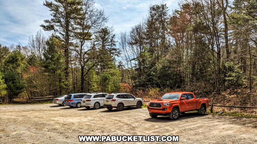 A gravel parking area at the trailhead of the Cliff Trail in Pike County, Pennsylvania, with several cars parked, indicating the popularity of the trail. A bright orange pickup truck stands out in the foreground, while the rest of the vehicles are more subdued in color. Tall pines and leafy trees surround the lot, offering a sense of seclusion and a clear starting point for adventurers seeking to explore the scenic overlooks above the Delaware River. The day is bright and sunny, casting sharp shadows on the ground and suggesting good weather for hikers.
