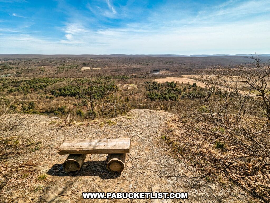 A wooden bench perched at the edge of a scenic overlook on the Cliffs Trail in Pike County, Pennsylvania. The bench sits on a gravelly area, offering a resting spot for hikers to enjoy the expansive view. The vista includes rolling hills, patches of forests and open fields, all under a wide, cloud-streaked blue sky. The Delaware River is not visible in this frame, but the landscape suggests a high vantage point above a river valley. The season appears to be early spring, with trees still bare and the grass just beginning to show green hues.