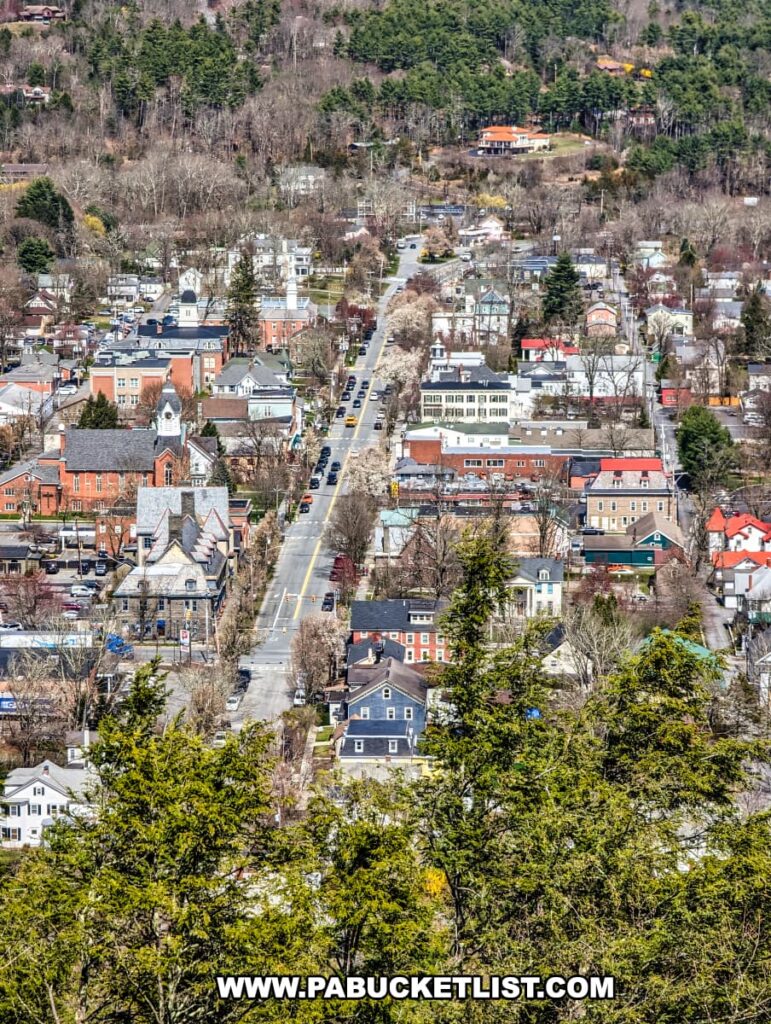 An view of downtown Milford from the Milford Knob Overlook on the Cliff Trail in Pike County, Pennsylvania. The image captures the town's quaint streets lined with a mix of residential homes and commercial buildings. A main street stretches through the center, with vehicles parked along the sides, leading the eye through the orderly grid of the town. The architecture displays a variety of colors and styles, characteristic of small-town charm. The surrounding landscape features patches of trees in the foreground, transitioning to the denser woodland that climbs the hills in the background, underlining the town's close relationship with the natural environment.