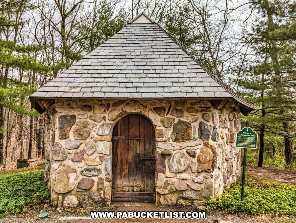 The photo showcases the quaint St. Columba's Chapel at Columcille Megalith Park, constructed of mixed stone with a heavy wooden door and wrought-iron hardware. The chapel's steeply pitched roof features weathered shingles, and it's complemented by a backdrop of dense woodland. A green informational sign stands to the right, identifying the structure and contributing to the park's rustic and historical character.