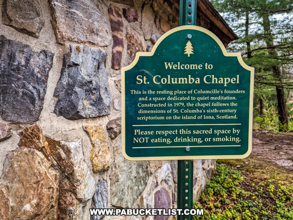 The image displays a close-up of a green informational sign next to the textured stone wall of St. Columba Chapel at Columcille Megalith Park. The sign welcomes visitors to the chapel, describing it as the resting place of the park's founders and a space dedicated to quiet meditation. It mentions that the chapel, constructed in 1979, is modeled after St. Columba's sixth-century scriptorium on the Isle of Iona, Scotland. The sign requests visitors to respect the sacred space by not eating, drinking, or smoking. Behind the sign, the natural setting of the park is partially visible, with trees and undergrowth contributing to the serene atmosphere.