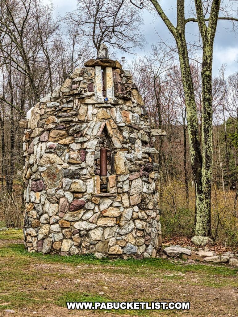 The image shows a distinctive, tall stone tower at Columcille Megalith Park, built with an assortment of rocks and boulders of various shapes and colors. The rustic tower features a wooden door and is topped with a small stone cross, giving it a historical and ecclesiastical appearance. Bare trees surround the tower, with the overcast sky above hinting at the quiet, contemplative nature of the park.