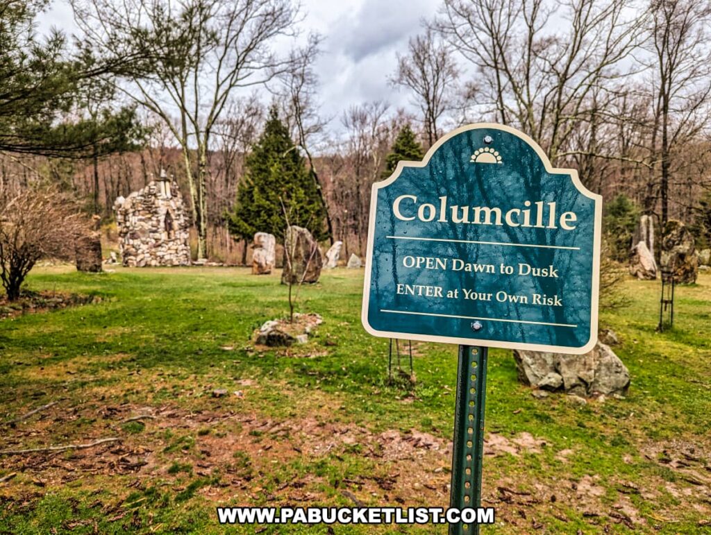 In the foreground of the photo is a sign at Columcille Megalith Park which reads "Columcille, OPEN Dawn to Dusk, ENTER at Your Own Risk" against a teal background with a semi-circular motif. Behind the sign, a meadow stretches towards a distinctive stone tower and various standing stones, reminiscent of ancient megaliths, scattered across a lush green landscape. The park's natural beauty is further accentuated by the backdrop of a cloudy sky and forested horizon, inviting exploration and reflection.