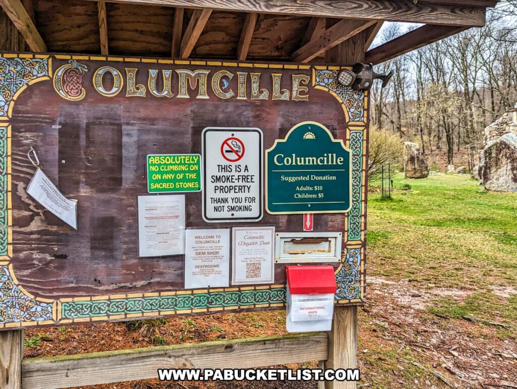 The photo shows an information kiosk at the entrance of Columcille Megalith Park. The kiosk is adorned with Celtic designs and provides various notices including an "ABSOLUTELY NO CLIMBING ON ANY OF THE SACRED STONES" warning, a "THIS IS A SMOKE-FREE PROPERTY" reminder, and a sign suggesting donations for entry. A red donation box and informational maps are available, with a clear view of the stone structures and wooded landscape of the park in the background. The scene is indicative of a well-maintained and respectfully presented historical site.