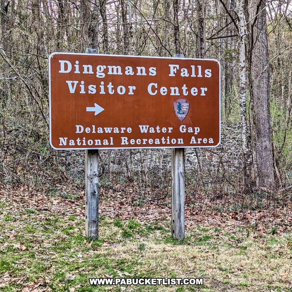 An informative sign stands prominently in a natural setting, its rusty red background catching the eye. It reads "Dingmans Falls Visitor Center" with an arrow pointing to the right, indicating the direction to proceed. Below, in bold letters, it says "Delaware Water Gap National Recreation Area," suggesting that the visitor center is part of this larger protected area. Leafless trees in the background suggest a late fall or early spring season, with the ground covered in a thin layer of brown leaves and the hint of green grass beginning to emerge. The scene captures the informative start of an adventure in Pike County, Pennsylvania.