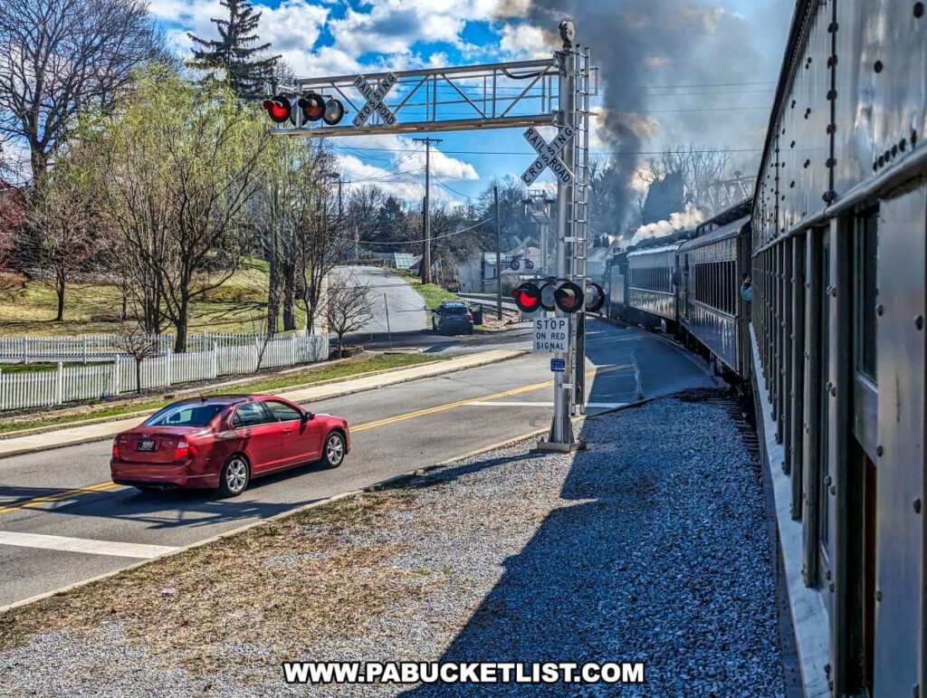 A classic steam train from the Everett Railroad is captured mid-journey as it crosses an intersection in Pennsylvania, with a red car paused at the crossing, awaiting its passage. The train's passenger cars, adorned with rivets and flush with historic character, follow closely behind the billowing smoke of the engine. The crossing signal, with its bright red lights and stop sign, stands prominently at the forefront, a reminder of the safety measures in place where rail meets road. Overhead, a clear blue sky provides a striking background to the scene, highlighting the contrast between the powerful, old-fashioned locomotive and the modern vehicle. This snapshot, taken during the "Spring into the Cove" excursion, encapsulates a moment where time-honored tradition intersects with the pace of contemporary life.