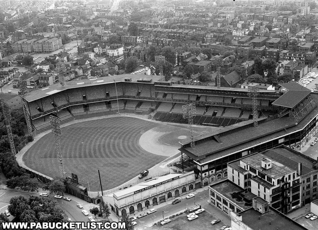 An aerial black and white photograph captures Forbes Field, the historic baseball park in the Oakland neighborhood of Pittsburgh, Pennsylvania. The stadium, with its distinctive double-decked grandstand and vast outfield, is set against a densely populated residential area. Cars from the era can be seen parked along the street, and the architecture of the surrounding buildings reflects the early to mid-20th century style. The ballpark is empty, creating a sense of stillness and anticipation, and the meticulous care of the diamond and field is evident even from this high vantage point. This image encapsulates the bygone days when Forbes Field was the heart of Pittsburgh's baseball history as the home of the Pirates.