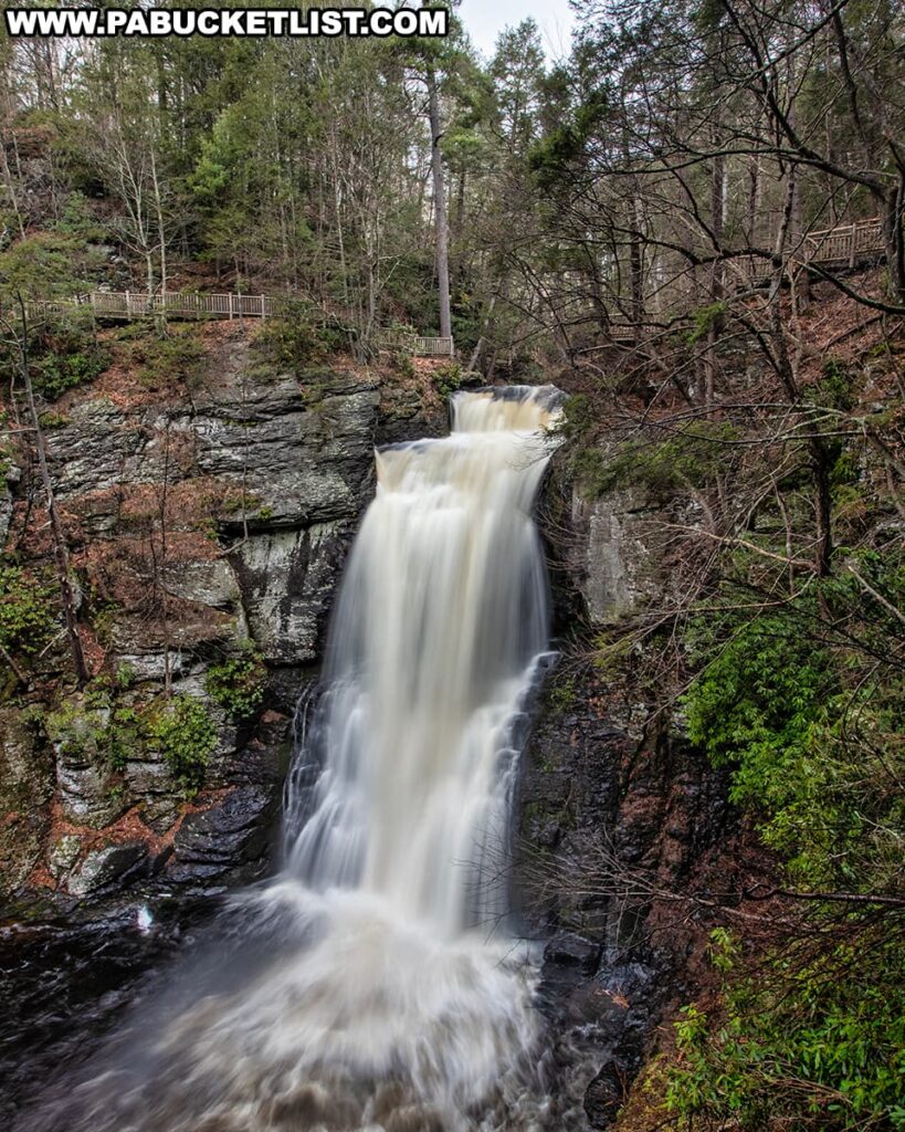 The main waterfall at Bushkill Falls in Pike County, Pennsylvania, cascades powerfully down a rocky cliff surrounded by the lush greenery of the Pocono Mountains. The waterfall is viewed from a distance, revealing its impressive height and the misty spray at its base. Wooden observation decks are visible on either side of the falls, providing visitors with safe and strategic viewpoints to witness this natural wonder. The scene captures the dynamic beauty and the serene ambiance of the park, which is renowned for its series of stunning waterfalls and the rustic charm of the well-maintained wooden boardwalks that grant access to these natural spectacles.
