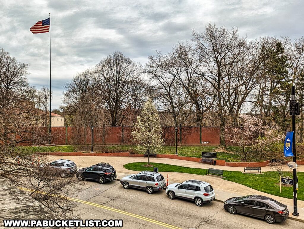 The photo offers a view from an elevated perspective, showcasing a section of the remaining outfield wall from the historic Forbes Field, with the distance marker "436 FT" visible on the brick surface. In the foreground, parked cars line the side of a street that borders a well-maintained grassy area with budding trees and a walkway, indicating the arrival of spring. An American flag flies proudly atop a tall flagpole, centered in the image. The setting is tranquil and residential, a stark contrast to the vibrant sports atmosphere that would have once filled the area when Forbes Field was the home of the Pittsburgh Pirates baseball team.