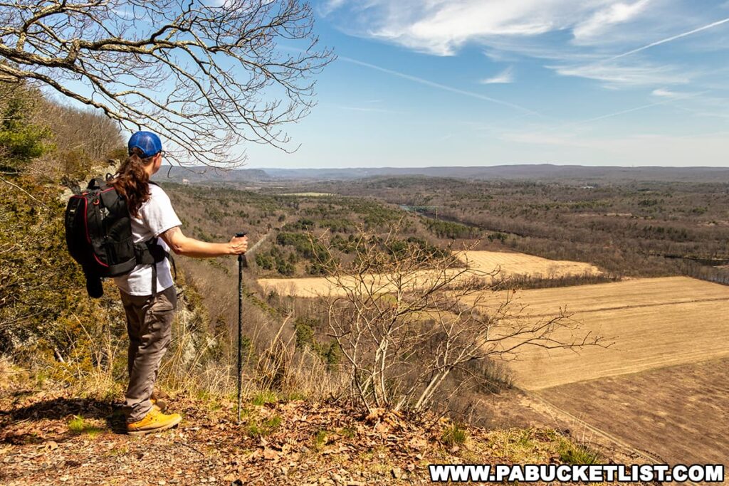 A hiker stands at the edge of a high cliff along the Cliff Trail in Pike County, Pennsylvania, admiring the expansive view. The trail is known for its scenic overlooks above the Delaware River. The hiker is wearing a white shirt, beige pants, and yellow hiking boots, with a blue cap and a black backpack. They're holding a walking stick in their right hand, gazing out towards the rolling hills and a patchwork of forest and fields stretching into the distance under a blue sky with wispy clouds. Early spring has left the trees bare, offering an unobstructed view of the landscape.