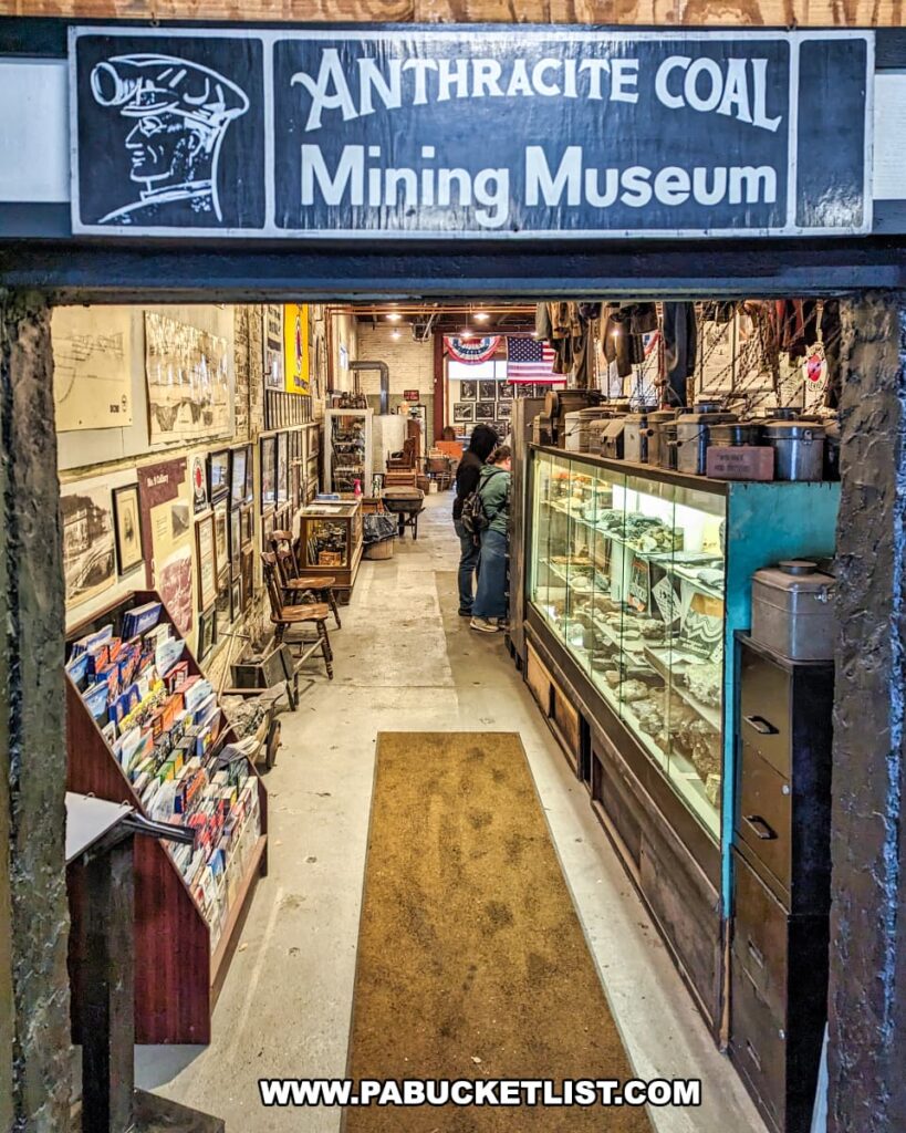 Entrance to the Anthracite Coal Mining Museum at the Number 9 Coal Mine in Carbon County, PA. A sign with bold lettering hangs above the doorway, depicting a miner's head in profile. Inside, visitors peruse exhibits along a narrow corridor lined with historical photographs, mining equipment, and memorabilia displayed in glass cases. The aisle is flanked by racks of postcards, ensuring that the mining heritage is not only preserved but also shared with others. The museum's rustic interior, with its stone walls and warm lighting, creates an inviting atmosphere that transports visitors back in time to the era of anthracite coal mining.