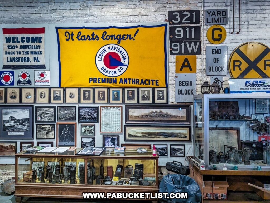 A wall of memorabilia at the Number 9 Coal Mine and Museum in Carbon County, PA, celebrates anthracite mining history. A large yellow banner proclaims "It lasts longer!" above the Old Company's Lehigh logo for "PREMIUM ANTHRACITE." Below, a variety of framed photographs, documents, and plaques line the wall, each telling a part of the coal mine's story. Railroad crossing signs, mining equipment, and a "WELCOME" banner for the 150th anniversary hang above glass cases displaying an array of antique mining lamps and tools. The exhibit, against a white brick backdrop, provides an immersive historical experience.