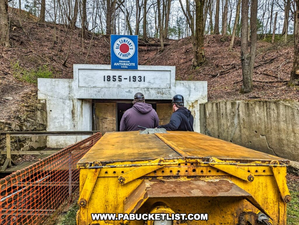 Two tour guides are about to enter the Number 9 Coal Mine in Carbon County, PA, seated on a yellow mine cart at the entrance. The mine entrance displays a sign above with the Old Company's Lehigh logo stating "PREMIUM ANTHRACITE COAL." The cart, positioned on tracks, is ready to descend into the tunnel, giving guests a genuine experience of a miner's journey underground. The surrounding area is a wooded hillside, with bare trees indicating the early spring season.