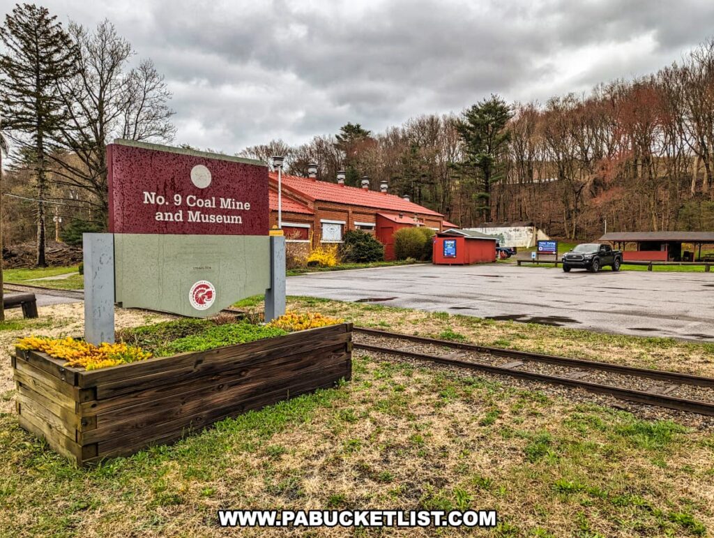 The entrance to the Number 9 Coal Mine and Museum with a maroon and gray sign surrounded by a wooden flower box filled with yellow flowers. The sign has the Old Company's Lehigh logo and announces the "No. 9 Coal Mine and Museum." In the background, a red building, likely part of the museum, sits beside an empty parking lot with a few cars and a blue portable restroom. Railroad tracks run in the foreground, hinting at the transport history associated with the mining industry.