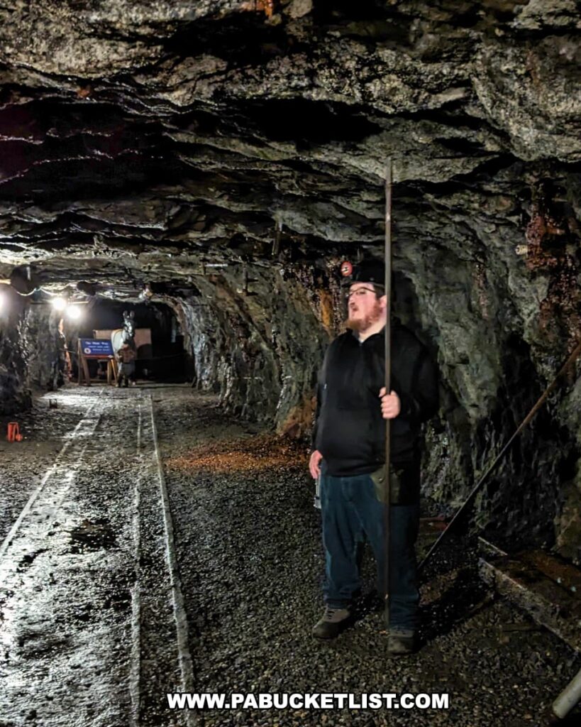 A tour guide, holding a long metal rod, stands inside the Number 9 Coal Mine in Carbon County, PA. He appears to be demonstrating or explaining a process as he gazes upward, likely discussing the mine's history or the mining process. The guide is wearing casual attire and a headlamp. The mine's tunnel surrounds him, with its dark, rugged rock walls and ceiling, illuminated by lighting that casts a glow on the scene. Further back in the tunnel, another person is visible near some equipment, possibly part of the tour group.