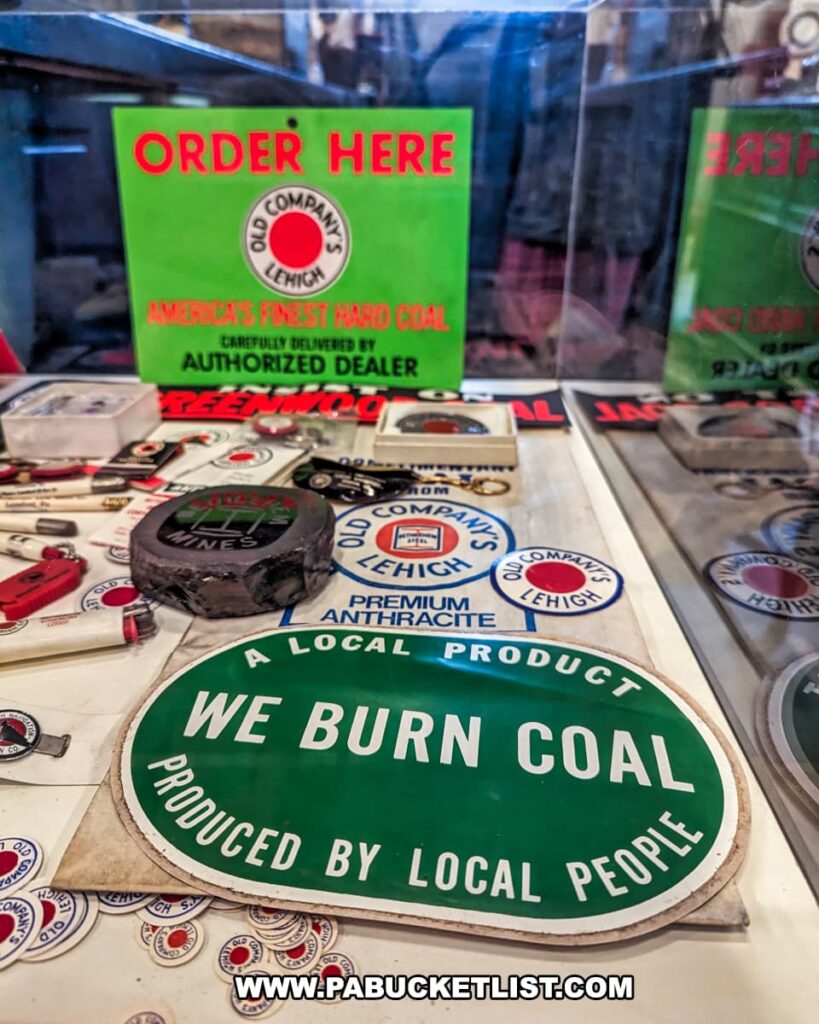 A display of coal mining memorabilia, including a bright green sign that reads "ORDER HERE" with the Old Company's Lehigh logo, stating "AMERICA'S FINEST HARD COAL" and "AUTHORIZED DEALER." Various badges and emblems with similar branding are scattered below. Prominently in the foreground is an oval-shaped sign with a green border and white center declaring "WE BURN COAL PRODUCED BY LOCAL PEOPLE." The items are showcased behind a transparent protective barrier.