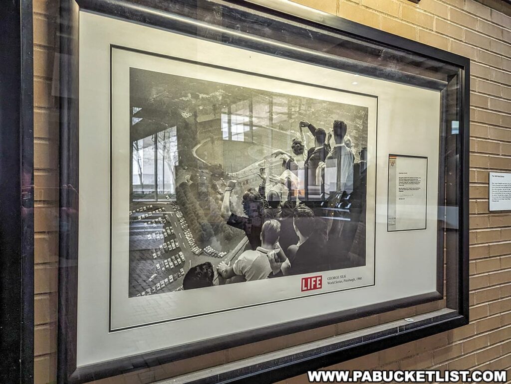 A framed photograph displayed on a brick wall, captured by George Silk for LIFE magazine, depicts a moment from the 1960 World Series at Forbes Field in Pittsburgh. The image shows a view of the stadium from a high vantage point, likely from the University of Pittsburgh's Cathedral of Learning, giving a glimpse into the excitement of the historic game. The photograph is part of an exhibit or collection, accompanied by descriptive text on the side, allowing viewers to connect with a significant event in the history of the Pittsburgh Pirates and their former home field.
