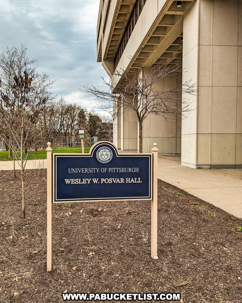 In the foreground stands a sign for the "University of Pittsburgh Wesley W. Posvar Hall," with the university's seal at the top. Behind the sign is a section of the building itself, a large structure with beige exterior walls and deep-set windows. This building stands on the grounds where Forbes Field once was, the historic baseball park and one-time home of the Pittsburgh Pirates. Leafless trees and mulched flower beds suggest it might be late fall or early spring. The photograph captures the legacy of Forbes Field as it is remembered and commemorated at the University of Pittsburgh.