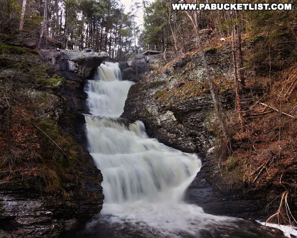 A serene depiction of Raymondskill Falls, Pennsylvania's tallest series of waterfalls, nestled in Pike County. Water flows powerfully over a series of rocky ledges, creating a multi-tiered cascade. The falls are framed by rugged cliffs and a variety of trees, some with bare branches and others with evergreen needles, indicative of the transition between seasons. A viewing deck with safety railings is perched discreetly at the top right, offering visitors a prime perspective of this natural spectacle. The surrounding forest floor is covered with a mix of fallen leaves and a dusting of snow, hinting at the cold weather.