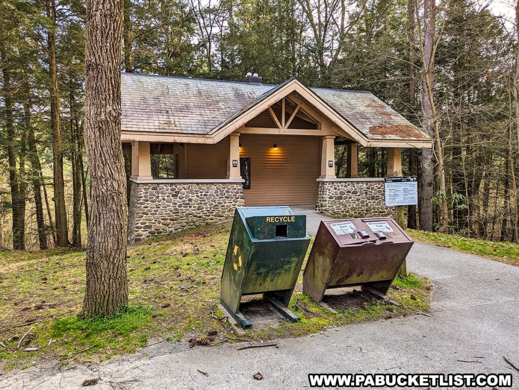 A restroom facility at the Raymondskill Falls parking area in Pike County, Pennsylvania, surrounded by a tranquil forest. The building features a stone foundation with a taupe facade and a gabled roof that has begun to show signs of moss and weathering. A single light fixture glows above the entrance, providing illumination as the daylight fades. In the foreground, there are two waste receptacles—one for recycling and one for trash—indicating a commitment to environmental stewardship. A bulletin board stands next to the building, likely providing visitors with information about the park and its facilities. Tall trees stand sentinel around the area, with the forest floor covered in grass and fallen pine needles.