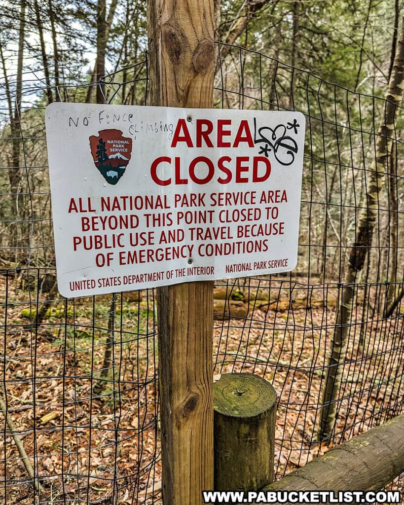A weathered sign attached to a wooden post with a wire fence in the background at Raymondskill Falls in Pike County, Pennsylvania, announces "AREA CLOSED." It states that all National Park Service areas beyond this point are closed to public use and travel due to emergency conditions, as declared by the United States Department of the Interior, National Park Service. The sign shows signs of wear and vandalism, including graffiti that reads "no fence climbing" and some stickers. It stands as a clear notice to visitors amidst a backdrop of leaf-covered ground and the bare branches of winter or early spring.