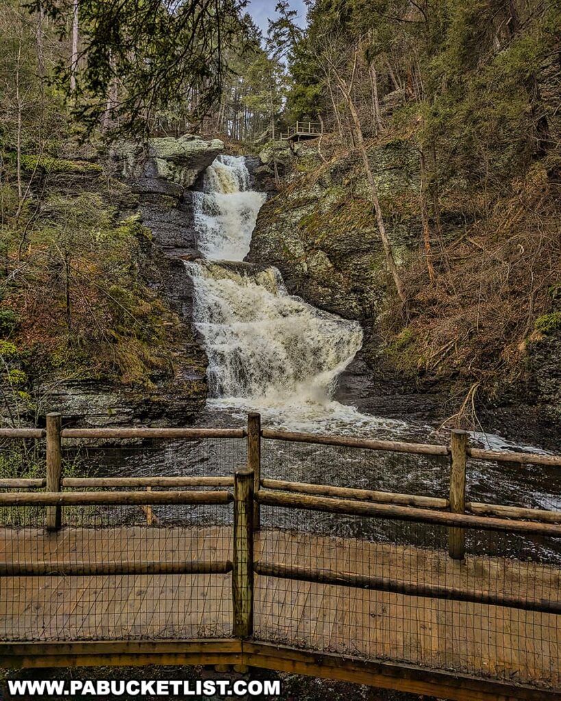 A viewing platform overlooks the lower section of Raymondskill Falls in Pike County, Pennsylvania. A wooden fence with wire mesh for safety encloses the platform. The falls cascade down in two main stages: a powerful drop from the upper level into a small pool and then a wider, frothy plunge to the base. Lush evergreen trees line the rocky edges, and a lookout with railings is visible above on the top right, suggesting multiple vantage points for visitors. The water's energy is palpable as it crashes into the pool below, creating a misty ambiance in the forested setting.
