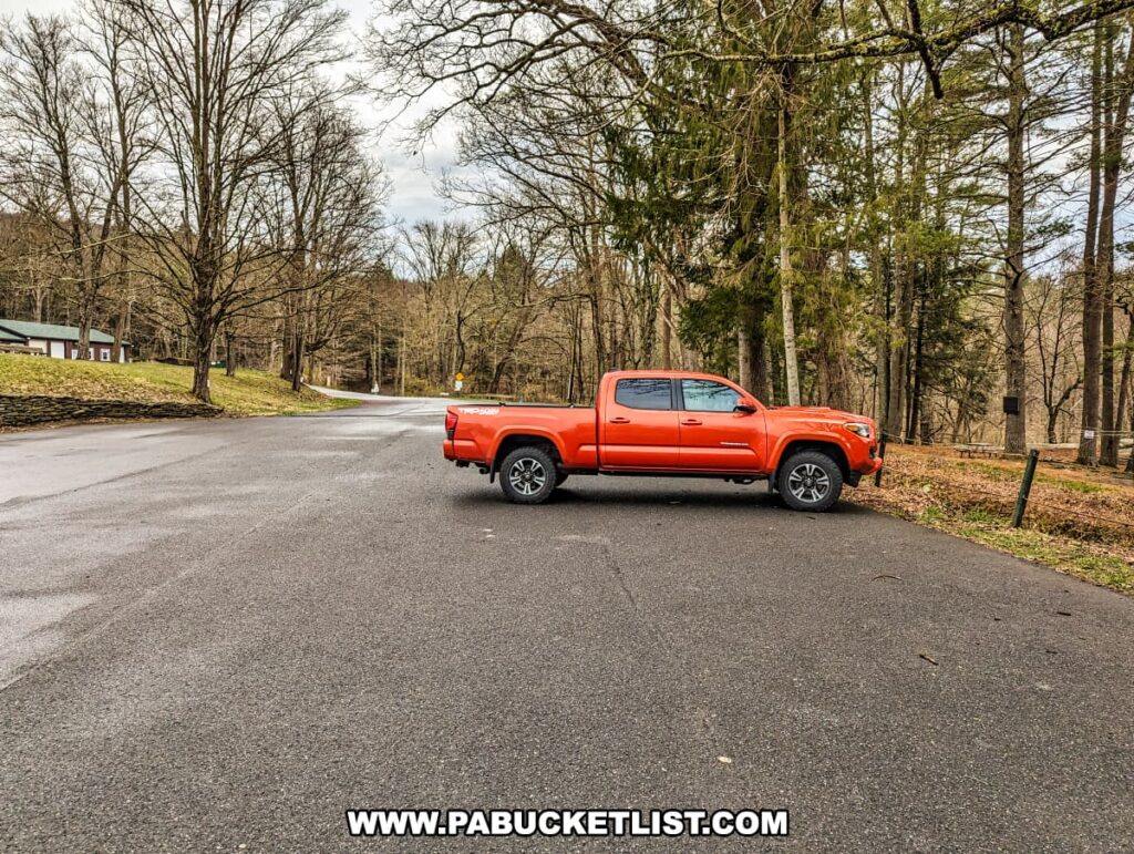 An orange pickup truck is parked on the side of a road that meanders through the Resica Falls Scout Reservation in Monroe County, Pennsylvania. The road is lined with bare deciduous trees and evergreens, suggesting a late fall or early spring season. A well-maintained grassy area with a wooden fence can be seen on the right, leading towards a forested area, indicative of the natural surroundings of the falls. The overcast sky above and the quiet road create a peaceful, rural landscape, just a short distance from the roadside attraction of Resica Falls.