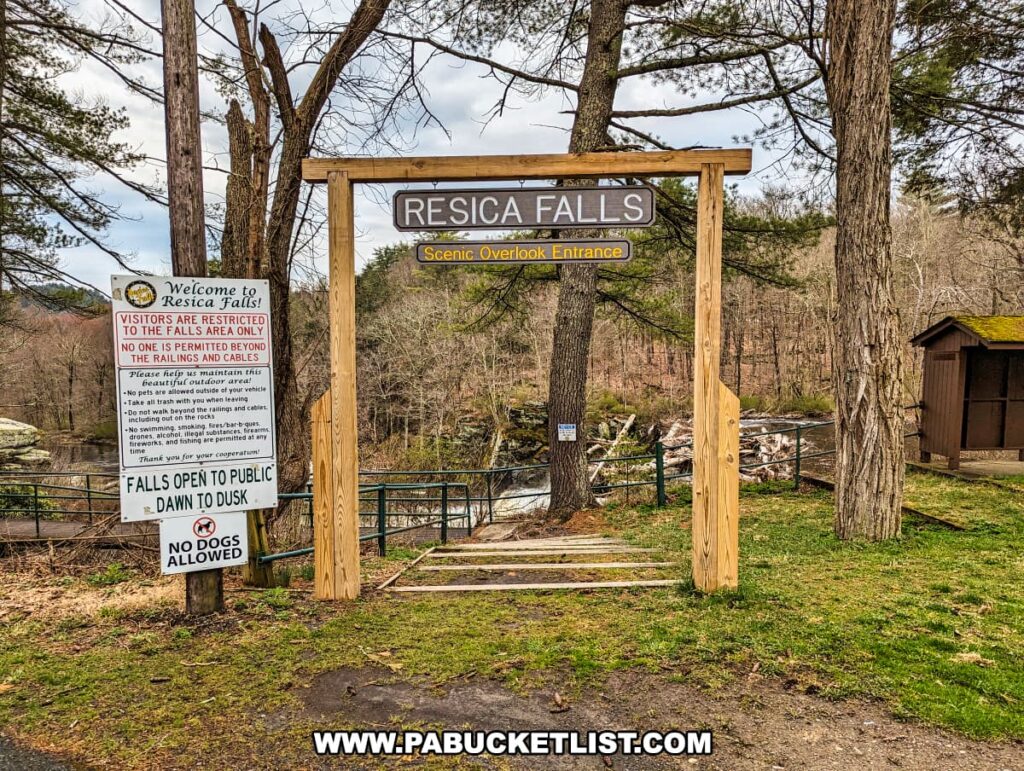 The entrance to the scenic overlook at Resica Falls is marked by a simple wooden gateway with a sign on top declaring "Resica Falls." A welcome notice is affixed to the left post detailing the rules and restrictions for visitors, emphasizing safety near the railings and cables, and noting that the area is open from dawn to dusk with no dogs allowed. Beyond the gateway, a glimpse of the waterfall can be seen through the trees, along with a rustic shed to the right, all surrounded by the forested landscape of the Scout Reservation. The path leading to the overlook promises a close view of the natural beauty the area is known for.