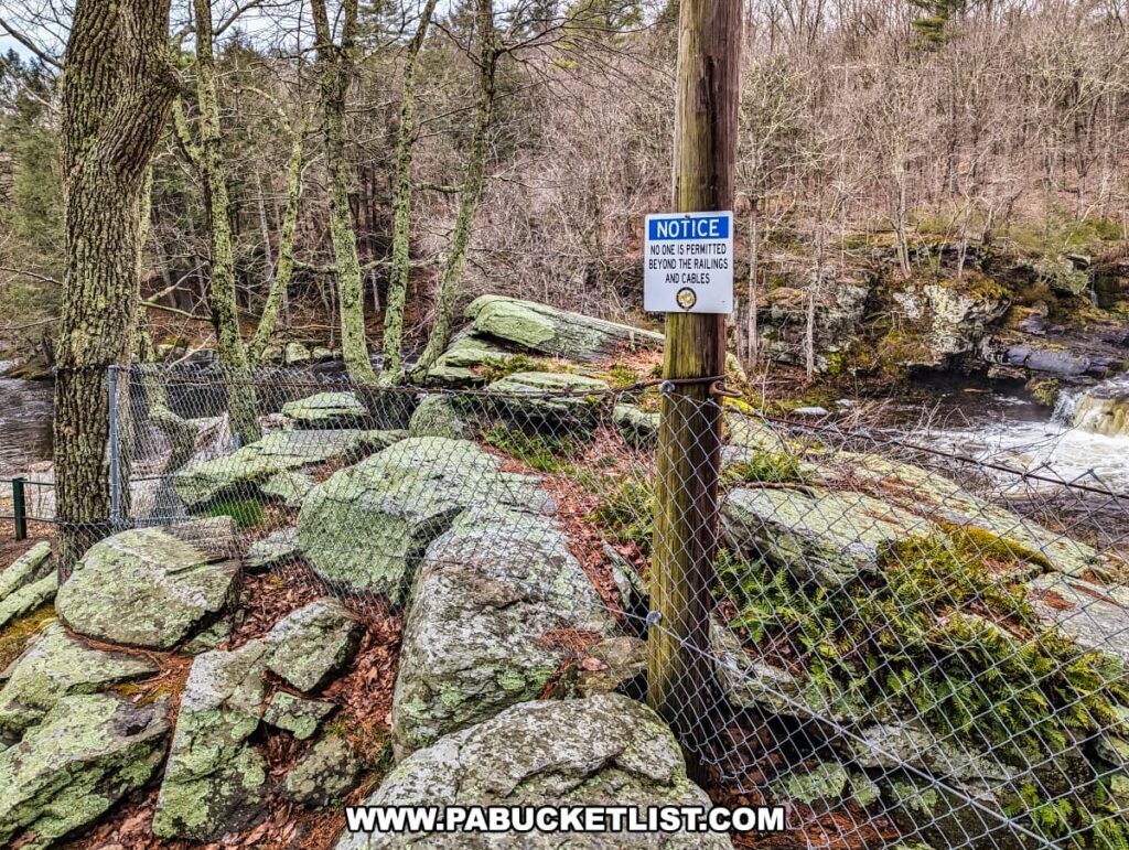 A safety notice sign is affixed to a wooden post next to a chain-link fence, warning that no one is permitted beyond the railings and cables at Resica Falls in Monroe County, Pennsylvania. The fence stands on a rocky outcrop covered in green moss, indicating the moist environment near the falls. Beyond the fence, the rushing waters of Resica Falls flow over rocks, surrounded by a forest of bare deciduous trees and evergreens. The natural scene is a mix of rugged wilderness and safety precautions, reflecting the blend of outdoor adventure and responsible stewardship promoted at the Resica Falls Scout Reservation.