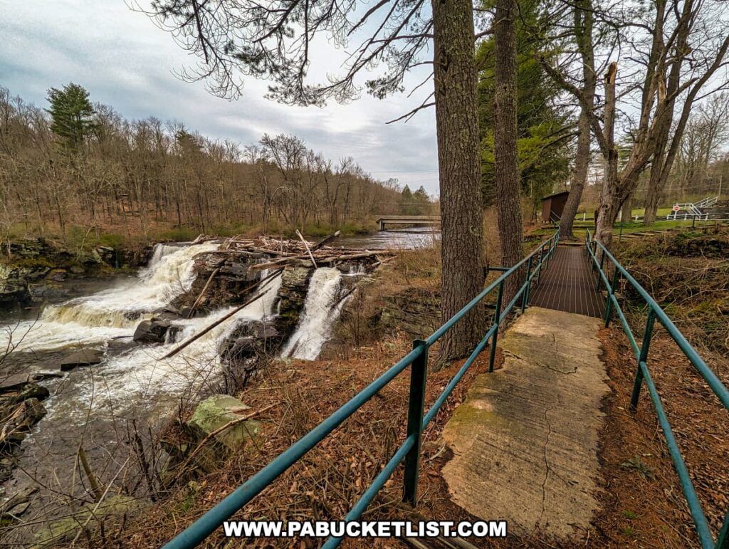 A sturdy walkway with teal railings guides visitors along the edge of the rushing Resica Falls in Monroe County, Pennsylvania. The path offers a close view of the waterfall, which is split into vibrant cascades tumbling over moss-covered rocks, with fallen trees adding a wild, natural element to the scene. To the right, the river stretches out calmly beyond the falls, with a simple bridge visible in the distance. The surrounding forest, in early spring regrowth, adds to the scenic beauty of the Resica Falls Scout Reservation, inviting exploration and reflection in this peaceful setting.