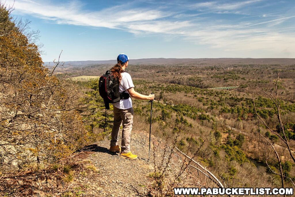 A hiker with long hair, wearing a blue cap, white shirt, and yellow boots, stands looking out over a scenic vista from the Cliff Trail in Pike County, Pennsylvania. They have a black backpack on and are holding a walking stick. The view overlooks a diverse landscape of forest, fields, and a winding section of the Delaware River. The horizon is dotted with hills and the sky is clear with few clouds. The foliage suggests it's either late spring or early fall, and the environment is tranquil and expansive.