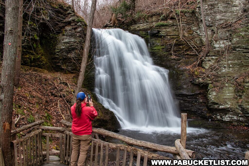 A visitor captures the beauty of Bridal Veil Falls at Bushkill Falls, Pike County, Pennsylvania, using a smartphone. The person, dressed in a red jacket and blue hat, stands on a rustic wooden observation platform that is part of the park’s extensive boardwalk system. The waterfall itself is a magnificent sheet of water cascading down a rock face, surrounded by the bare branches of trees in an early spring setting. This tranquil moment at the "Niagara of Pennsylvania" is emblematic of the park’s appeal, offering accessible, up-close encounters with nature’s splendor within the Pocono Mountains.