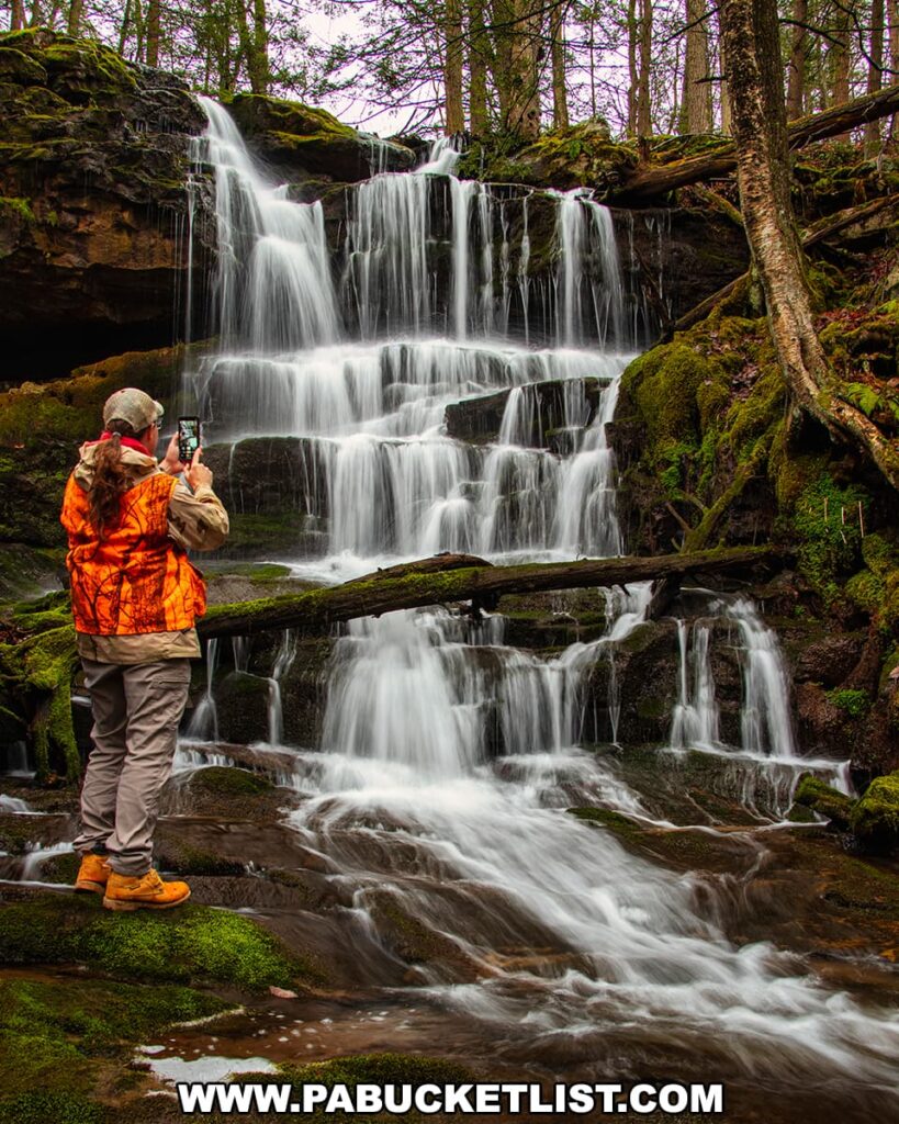 Author and photographer Rusty Glessner in outdoor attire, with a blaze orange vest and beige pants, stands on moss-covered rocks while photographing the captivating Salvatore Falls. The waterfall gracefully cascades over tiered rock formations, enveloped by the lush forest of State Game Lands 91 in Luzerne County, Pennsylvania. The surrounding woods are rich with moss and ferns, enhancing the natural beauty of this tranquil setting. The observer's presence adds a sense of scale and the human experience of nature's wonders.