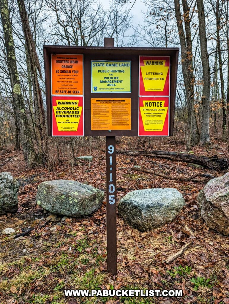 A signboard near Salvatore Falls on State Game Lands 91 in Luzerne County, Pennsylvania, displaying multiple notices. The signboard, set against a backdrop of barren trees, informs visitors of regulations including wearing orange during hunting seasons, the prohibition of alcoholic beverages and littering, and camping restrictions. Large boulders are scattered around the signboard, which is situated on a patch of grass with some leaf cover, indicative of autumn or early spring. A brown post in front of the sign displays an identification number "91105" in white lettering.
