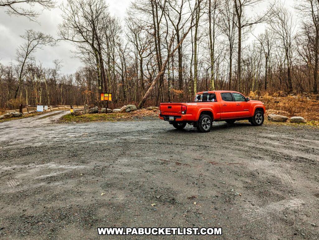 A parking area at Salvatore Falls on State Game Lands 91 in Luzerne County, Pennsylvania, featuring a vivid orange pickup truck parked on the gravel. Leafless trees surround the lot, suggesting a late fall or winter season. The sky is overcast, and the ground appears wet, possibly from recent rain. In the background, a gate blocks a road, with information signs posted nearby, inviting further exploration into the natural area. The landscape is stark, with the truck providing a stark contrast to the muted colors of the environment.